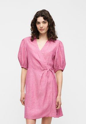 Γυναίκα με σγουρά σκουρόχρωμα μαλλιά, ντυμένη με ροζ κοντομάνικο φόρεμα τύπου wrap με φουσκωτά μανίκια, στέκεται μπροστά σε απλό φωτεινό φόντο.