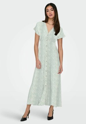 Kobieta ubrana w jasną, zieloną, krótkorękawową, zapinaną na guziki suknię maxi z delikatnym wzorem liści, stojąca nieruchomo w czarnych szpilkach.