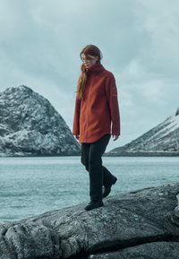 Rød fleecejakke med høj krave, prydet med et sidepatch, parret med sorte bukser og sort fodtøj, sat mod en klippet kystlandskab.