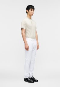 Πολό μπλουζάκι σε μπεζ με σχέδιο, με κοντά μανίκια, συνδυασμένο με λευκό παντελόνι στενής γραμμής και μαύρα μοκασίνια. Το μοντέλο στέκεται προφίλ.