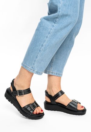 Feet wearing black platform sandals with buckle straps and silver studs, paired with light blue cropped jeans.