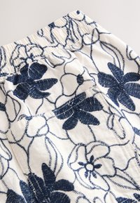 Blumengemusteter Stoff in Weiß mit blauer Stickerei; verfügt über einen gerafften Bund und ein kleines Taschen-Detail.