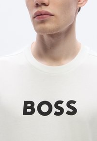 Valge pusa, mille esiküljel on suur must "BOSS" logo. Pehme, tekstuuriga kangas ja klassikalise ümarkaelusega.