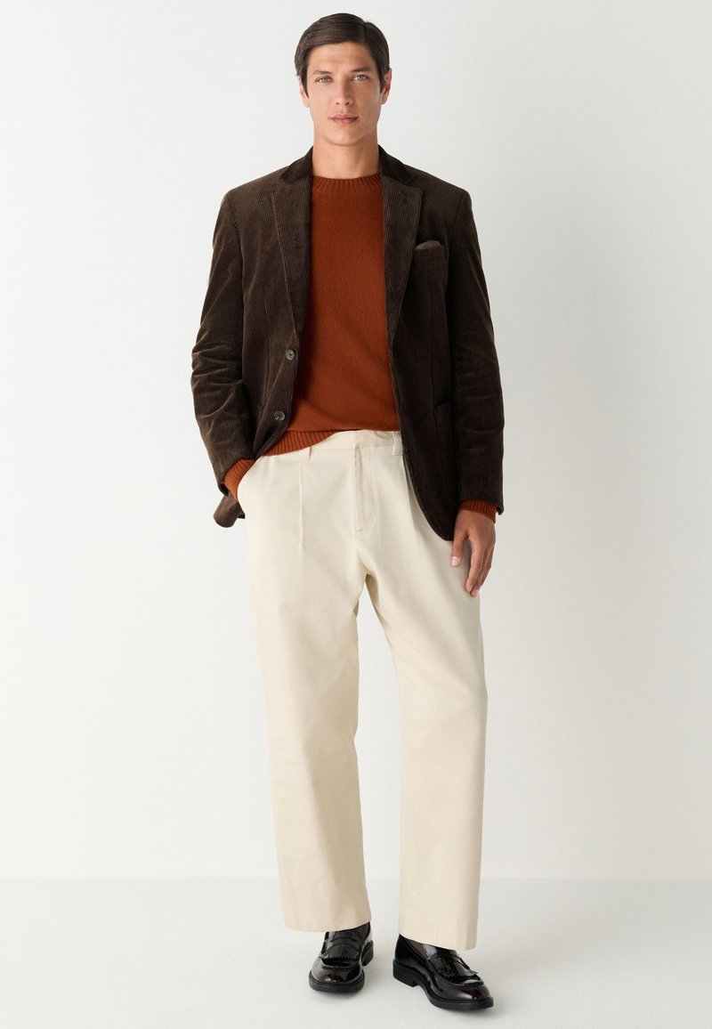 Blazer en velours côtelé marron porté sur un pull orange, accompagné d'un pantalon beige et de chaussures habillées noires. Le vêtement présente un col cranté et des poches.