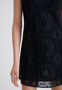 Vestido negro de encaje con patrón floral, que presenta recortes circulares y un dobladillo con forma de onda. La tela tiene un diseño texturizado e intrincado.
