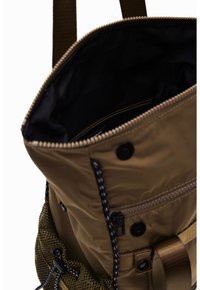 Beżowy plecak z zamkiem na górze, czarną podszyciem wewnętrznym, zewnętrzną kieszenią z siateczkową boczną ścianką oraz regulowanymi paskami.