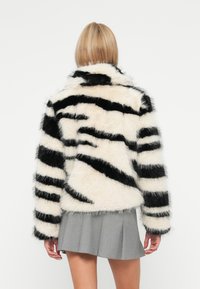 Lažni krzneni plašč z črnimi in kremasto belimi zebra črtami, z visokim ovratnikom in dolgimi rokavi, v kombinaciji s sivo naborkano krilom.