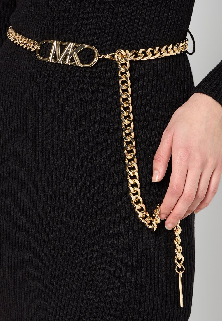 Cintura in metallo dorato con un design a catena chunky, dotata di una fibbia con logo e un lungo accento a catena, indossata sopra un capo nero a coste.