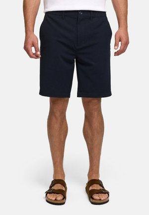 Homme portant un short noir jusqu'au genou, une chemise blanche et des sandales marron à bout ouvert, debout devant un fond uni.