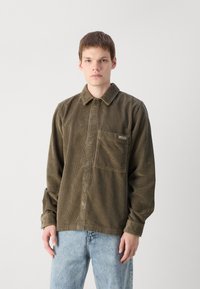 Olive green corduroy shirt with a button-down collar, front pocket, and a relaxed fit, paired with light blue jeans.