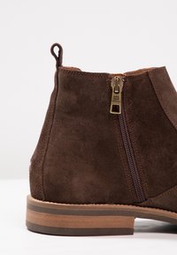 Brun mockaskor med ankellängd, med sidoclosure, dragflik vid hälen och staplad träledig på vit bakgrund.