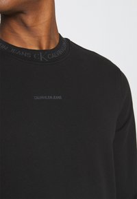 Sweat-shirt noir à manches longues avec un col côtelé, présentant un logo discret "Calvin Klein Jeans" sur la poitrine et une texture lisse.