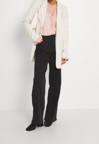 Žena v krémovém blejzru přes růžový volánkový svetr, černých kalhotách s vysokým pasem a širokými nohavicemi a černých kožených botách na podpatku, držící krémovou kabelku.