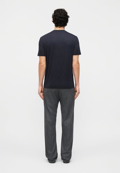 Άνδρας που στέκεται με την πλάτη, φοράει απλό μαύρο κοντομάνικο t-shirt και φαρδιά γκρίζα παντελόνια σε λευκό φόντο.