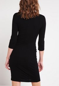 Robe noire ajustée à manches trois-quarts, avec une texture lisse et un design flatteur qui souligne la taille et les hanches.