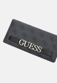 Billetera negra con exterior texturizado que presenta un patrón sutil de logotipos. Acentos de hardware en plata "GUESS" en una banda negra lisa. Forma rectangular.