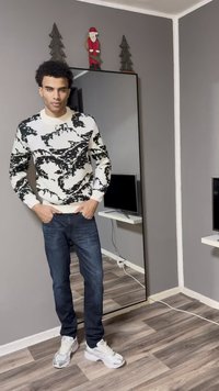 Maglione con motivo bianco e nero, jeans blu scuro e sneakers bianche. In piedi davanti a una parete grigia con uno specchio e una TV sullo sfondo.