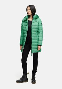Groene pufferjack met capuchon, gewatteerd ontwerp en voorrits. Draagt over een zwarte top en gecombineerd met zwarte broek en laarzen.