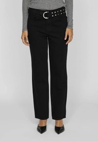 Jeans negros de pierna recta con un cinturón con tachuelas y hebilla plateada, combinados con zapatos negros de punta y una camiseta gris de manga larga.