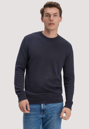 Mann trägt einen marineblauen Rundhalspullover und blaue Jeans und steht vor einem einfarbigen hellgrauen Hintergrund.