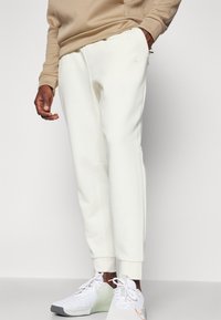 White sweatpants made of soft material, featuring a tapered leg design and an elastic waistband. Paired with white shoes, showcasing contrasting accents.