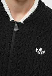 Čierna pletená bunda so vzorom z pleteného lana, rebrovaný biely golier, zips na prednej strane a malé biele logo Adidas na ľavej strane.