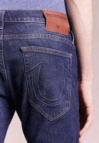 Mörkblå True Religion-jeans på en person, med fokus på bakfickan som har signaturhästsko-stickning och lädermärke på midjebandet.