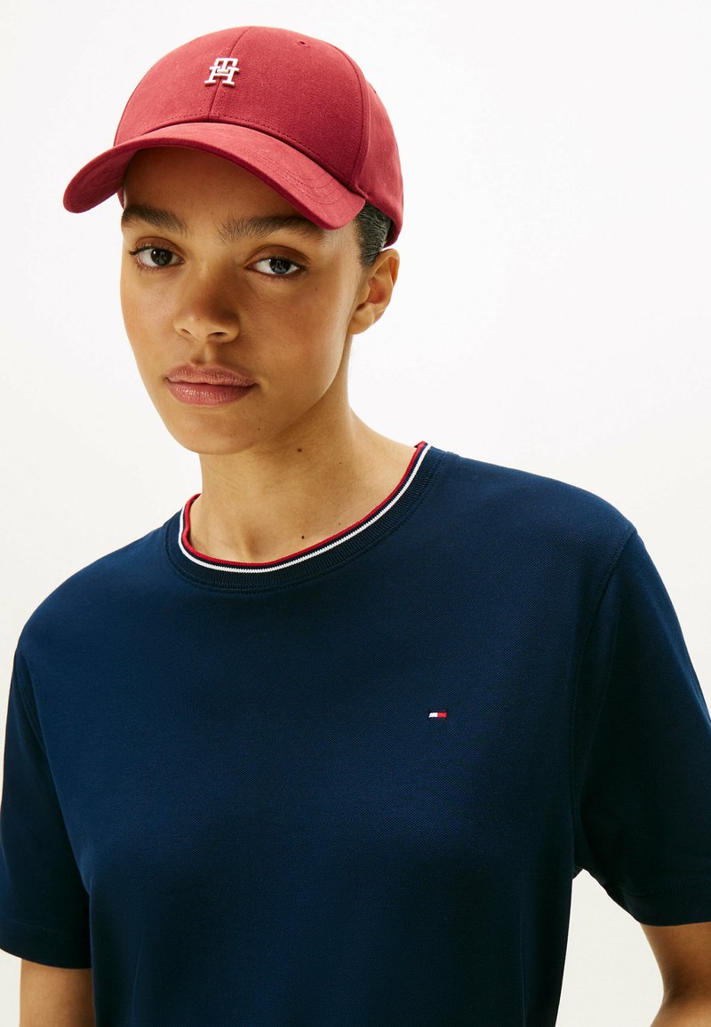 Rote Baseballmütze mit Logo, marineblaues T-Shirt aus strukturiertem Stoff mit geripptem Kragen und roten sowie weißen Akzenten. Nahaufnahme.