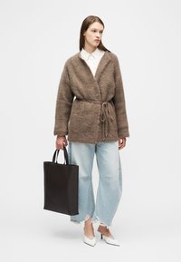 Γυναίκα που φοράει καφέ ζεστή ζακέτα με ζώνη, λευκό πουκάμισο με γιακά, φαρδιά σκισμένα ανοιχτό μπλε τζιν, άσπρα τακούνια, κρατώντας μια μεγάλη μαύρη τσάντα ώμου.