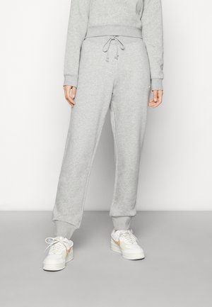 Personne portant un pantalon de survêtement gris clair avec une taille à cordon et des baskets blanches, debout devant un fond uni.