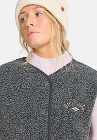 Grijze fleece vest met een textuurafwerking, voorzien van donkere randen en een klein geborduurd logo. Draag over een lichtroze gebreide kraag.