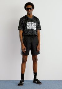 Black Adidas jersey with white graphic print, black shorts, and black Adidas footwear. Model also wears sunglasses and black socks.