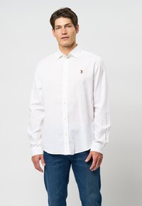 White long-sleeve button-up shirt made of linen fabric, featuring a small embroidered logo on the chest. Worn with blue denim jeans.