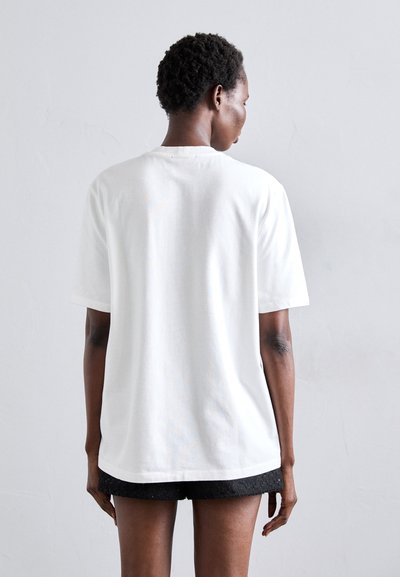 T-shirt en coton blanc à manches courtes et col rond, avec une coupe décontractée et des détails de couture visibles, associée à un short noir texturé.