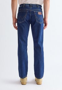 Personne portant un jean en denim bleu avec des poches arrière et des chaussures marron clair, debout devant un fond clair uni.
