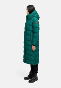 Cappotto lungo in teal imbottito con cappuccio, caratterizzato da un design trapuntato e tasche laterali con zip, abbinato a pantaloni neri e stivali con lacci.