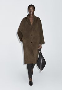 Brun uldfrakke med slagkrave, dobbeltbrystet design og sorte knapper. Sammen med sorte bukser og en lædertaske.