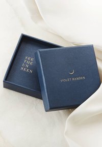 Eine marineblaue Box mit einer glatten Textur, die goldene Schriftzüge "SEE THE UNSEEN" und "VIOLET HAMDEN" sowie ein Halbmondakzent auf dem Deckel aufweist.