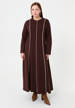 Donna con capelli castani lisci, che indossa un lungo vestito marrone con strisce verticali bianche e stivaletti marroni, in piedi davanti a uno sfondo bianco.