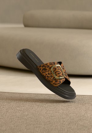 Sandale mule avec bride imprimé léopard, boucle dorée et semelle noire flottant au-dessus d'un tapis beige texturé.