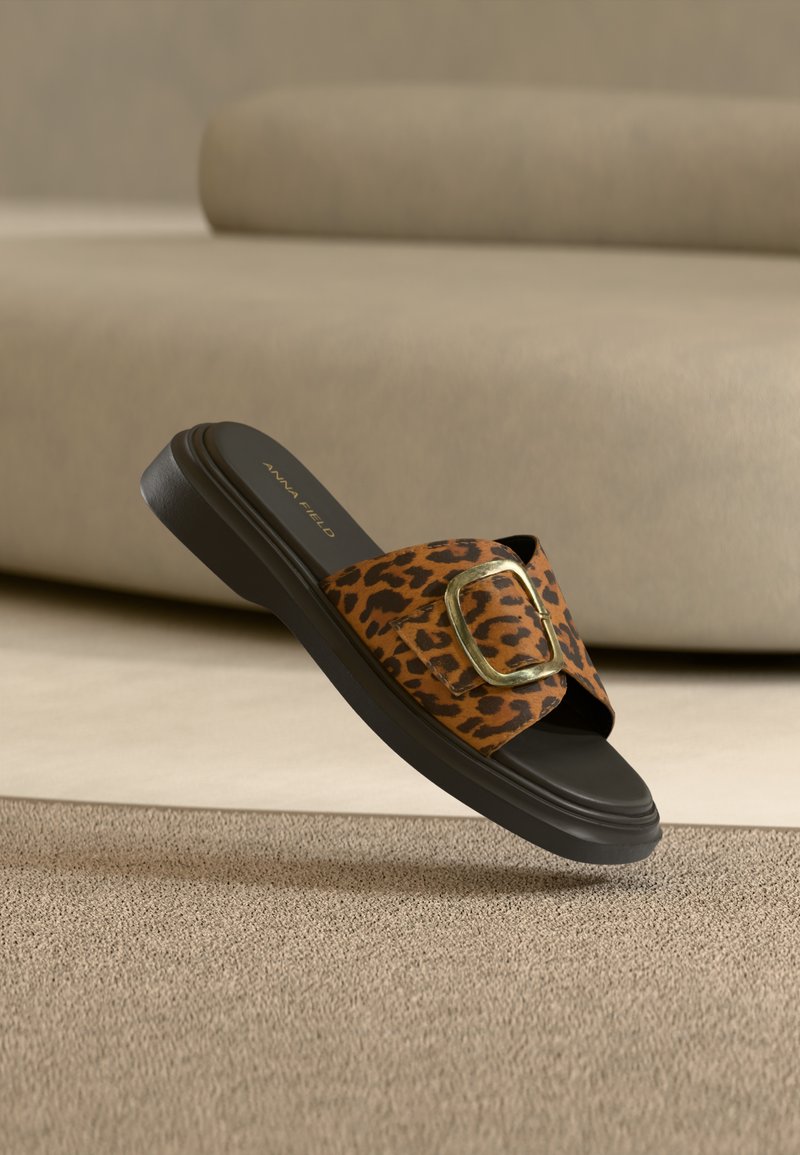 Σανδάλι τύπου σλάιντ με λουράκι σε σχέδιο λεοπάρδαλης, χρυσή αγκράφα και μαύρη σόλα, να αιωρείται πάνω από υφασμένο μπεζ χαλί.
