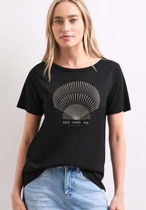 Ung kvinde med blondt hår iført en sort t-shirt med en solstrålegrafik og ordene "Sun Sand Sea" kombineret med lyseblå jeans.