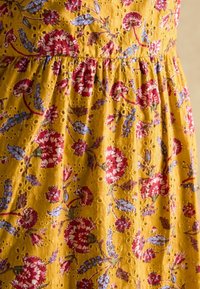 Kollane kangas, millel on punaste ja siniste õiekoosluste tikand. Omab auke ja volangiga vöökohta tekstuuri ja kujunduse lisamiseks.