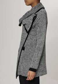 Abrigo de punto con un alto cuello y gran solapa delantera; esquema de color en blanco y negro, con detalles de puños y dobladillo acanalados.