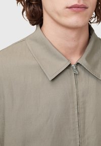 Lys olivengrøn skjorte med klassisk krave og frontlynlås. Stoffet har en struktureret overflade og har pæne syninger langs kanterne.