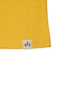 Gul t-shirt i bomull med en vit tygetikett som har svarta korsade bestick och texten "BXR". Mjuk textur med sydda kanter.