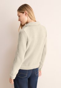 Giacca in pile grigio chiaro con collo a rivetto, maniche lunghe e finitura texturizzata. Presenta una vestibilità aderente e dettagli a orlo grezzo.