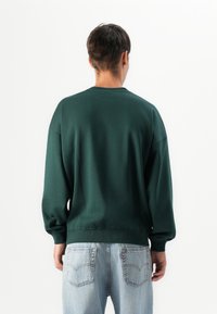 Man facing away wearing a dark green sweatshirt and light blue jeans against a plain white background.