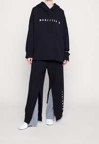 Felpa nera over size con testo "MARGIELA 6", abbinata a pantaloni felpati neri e grigi con spacchi laterali e scarpe bianche.