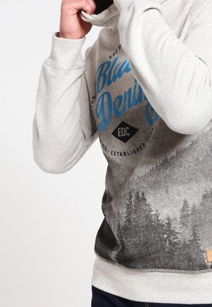 Personne ajustant le col d'un sweat-shirt gris clair avec le texte bleu "Blue Denim" et un graphisme de forêt sur la moitié inférieure.
